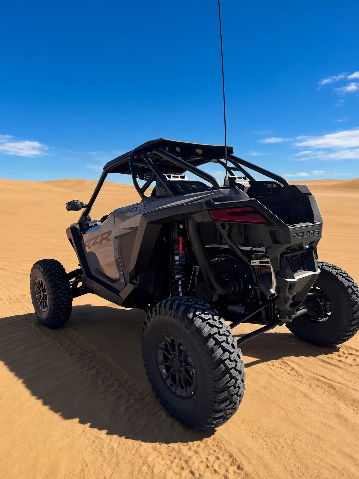 Polaris RZR Pro S Ultimate "Ninja" on bright sand dunes under a deep blue sky, rear three-quarter view showing its roll cage and red-reservoir shocks